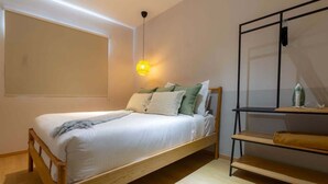 Ein Einzelbett mit Holzrahmen und weißer Bettwäsche, ein kleines Regal mit Spiegel sowie eine wandmontierte Leuchte.