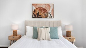 Una cama bien hecha con un edredón blanco, flanqueada por dos mesitas de noche con lámparas y una cesta tejida en cada una. Un cuadro abstracto de gran tamaño cuelga sobre la cama.