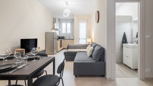 Ein modernes Wohnzimmer mit Essbereich, einem grauen Sofa, einem Fernseher und einer Küche im Hintergrund.