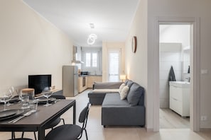 A modern living room with a dining area, a grey sofa, a TV, and a kitchen in the background. - Maison Rosaria Vacation Home near Allianz Stadium with Balcony (Torino)