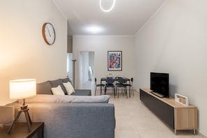 Uma sala de estar moderna com sofá, televisão sobre um console de madeira, mesa de jantar com cadeiras, relógio na parede e uma pintura.