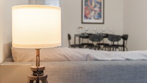 Ein Bett mit grauer Decke, eine Nachttischlampe und ein Essbereich im Hintergrund.