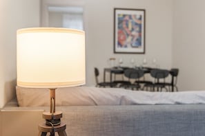 A bed with a grey blanket, a bedside lamp, and a dining area in the background. - Maison Rosaria Vacation Home near Allianz Stadium with Balcony (Torino)