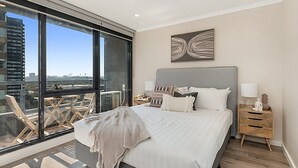 Une chambre moderne avec un grand lit, une table de chevet, une table en verre avec des chaises et une vue sur la ville.