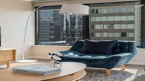 Un salon moderne avec un canapé bleu foncé, une table basse ronde, un lampadaire et une vue sur de hauts immeubles par une grande fenêtre.