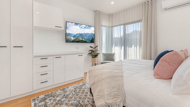 Une chambre moderne avec un lit, un téléviseur à écran plat fixé au mur, une plante en pot et une vue sur les montagnes par la fenêtre.
