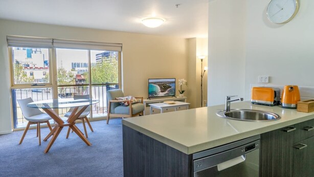 A modern kitchen with a large window, a dining area with a glass table, and a view of buildings outside. - Spacious City Stay Near Lygon Street Dining (Carlton)