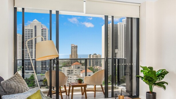 A modern living room with a large window, a sofa, two chairs, a table, and a potted plant. - 2-Bed Beach Apartment with Pool, Parking & Balcony (Surfers Paradise)