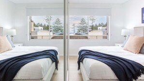 A mirrored room with two beds, a large window, and a view of trees and buildings. - MadeComfy Trendy Apartment on Dee Why Beach (Dee Why)