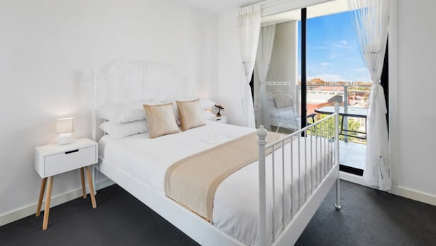 A modern bedroom with a white bed, a bedside table, and a view of the outdoors through a large window. - Executive and Spacious Apartment with Balcony (Sunshine)