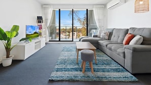 A modern living room with a grey sofa, a coffee table, a TV on a white stand, a potted plant, and a view through a glass door. - Executive and Spacious Apartment with Balcony (Sunshine)