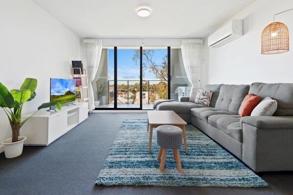 Una sala de estar moderna con un sofá gris, una mesa de centro, un televisor sobre un mueble blanco, una planta en maceta y vistas a través de un ventanal.