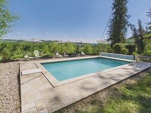 A rectangular swimming pool with a stone deck, surrounded by green chairs and umbrellas, set against a backdrop of trees and hills. - Manor with Terrace and Pool for 12 Guests (Saint-Léon-sur-Vézère)