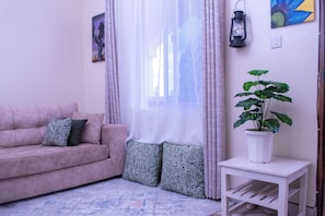 Ein Wohnzimmer mit einer Couch, gemusterten Kissen, einem Blumentopf, einem kleinen Tisch und einem Fenster mit durchsichtigen Vorhängen.