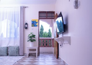 Ein Zimmer mit Schachbrettboden, einer Zimmerpflanze, einem an der Wand befestigten Fernseher und einem Fenster mit Blick auf Bäume.