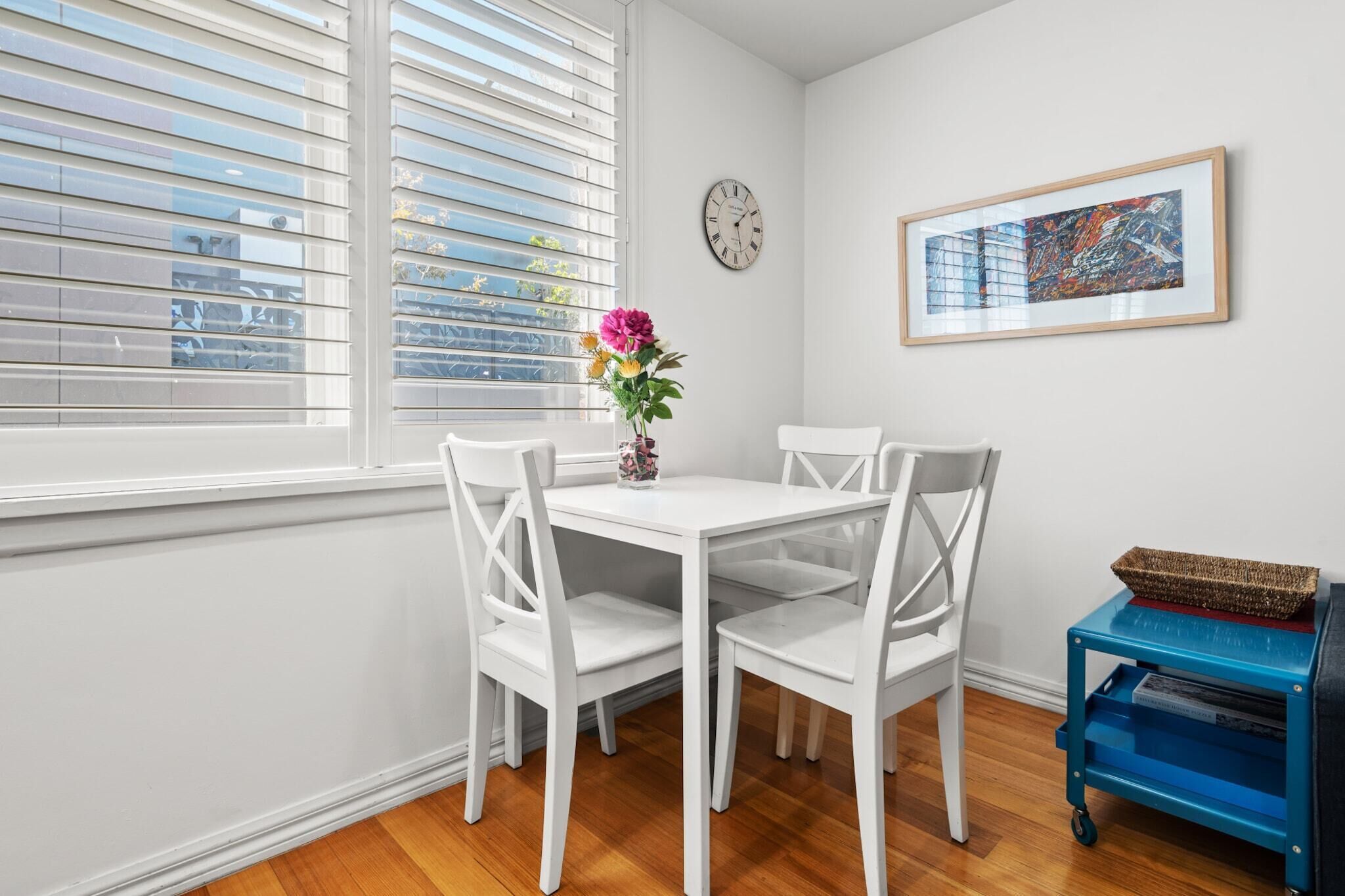 A dining area with a white table and chairs, a vase with flowers, a clock, and a framed artwork on the wall.
