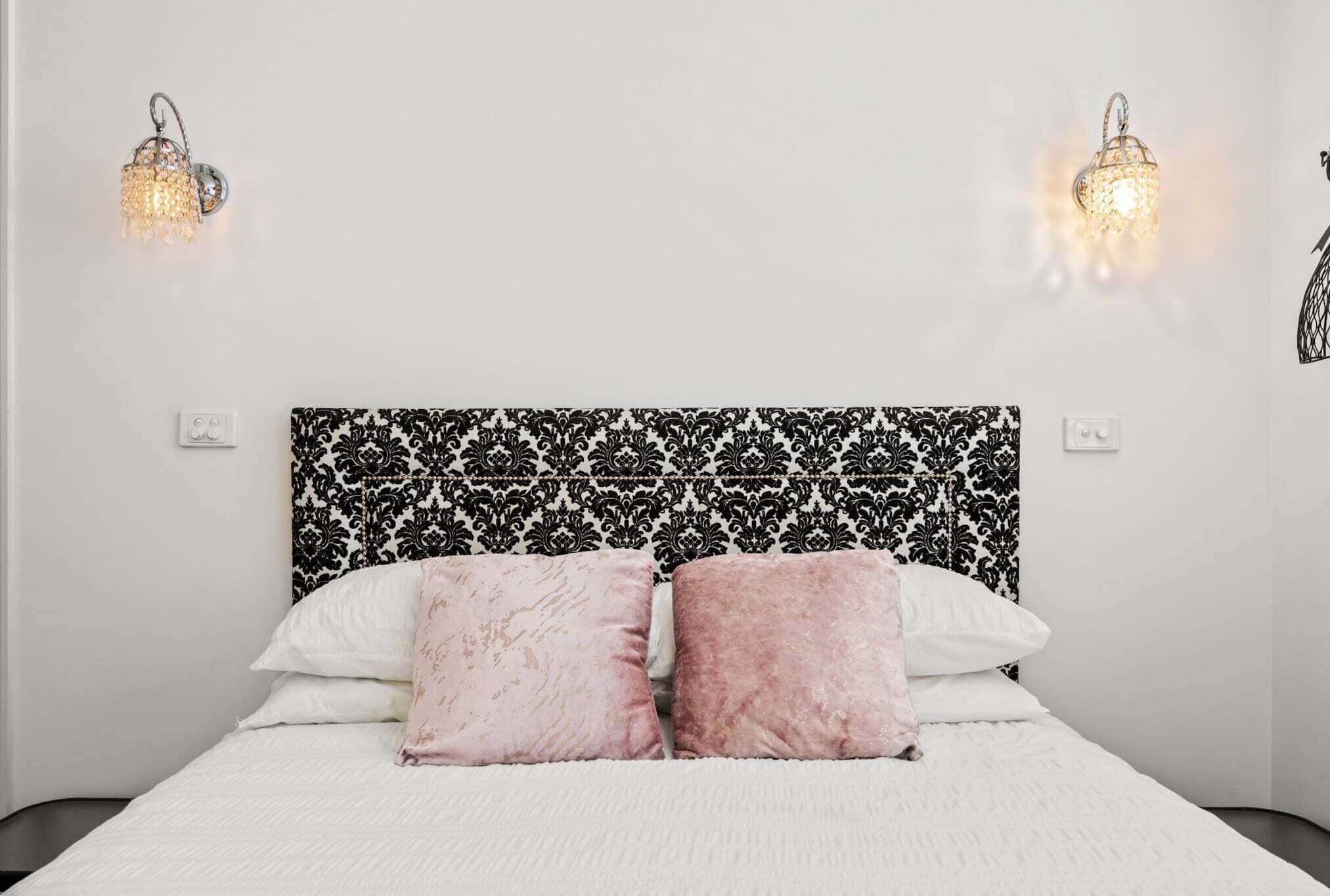 Un lit avec une tête de lit à motifs, deux appliques murales et un mur blanc recouvert d’un papier peint noir et blanc à motifs.
