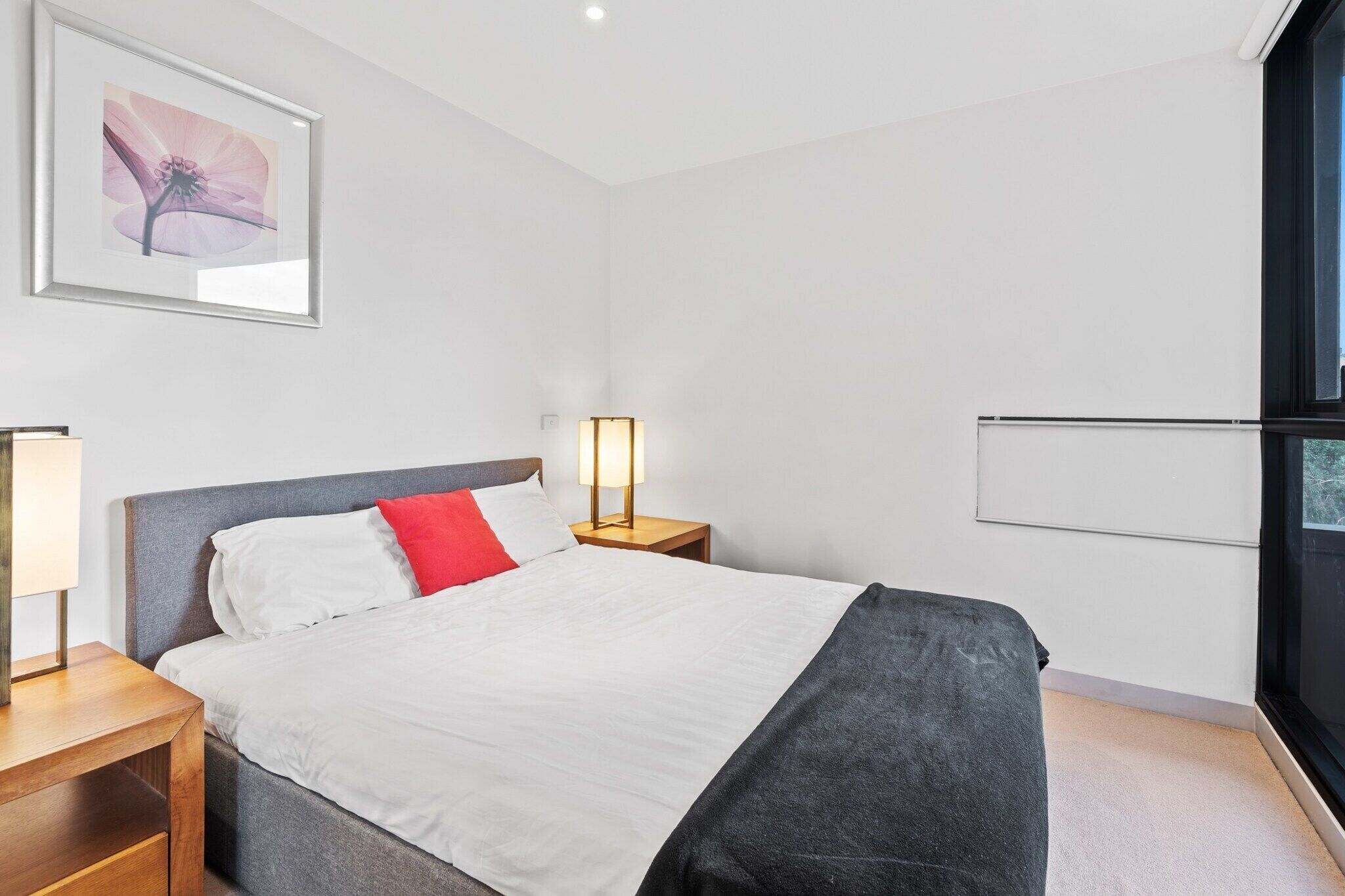 A modern hotel room with a large bed, a bedside table, a nightstand, a framed artwork on the wall, and a window with a view.