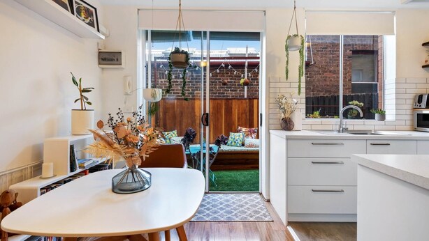 Une cuisine moderne avec des meubles blancs, un coin repas avec une table et des chaises blanches, et une vue sur un mur de briques et de la verdure à l’extérieur.