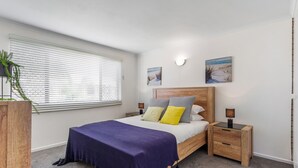 Un dormitorio con cama, mesitas de noche, persianas en la ventana y decoración en la pared.