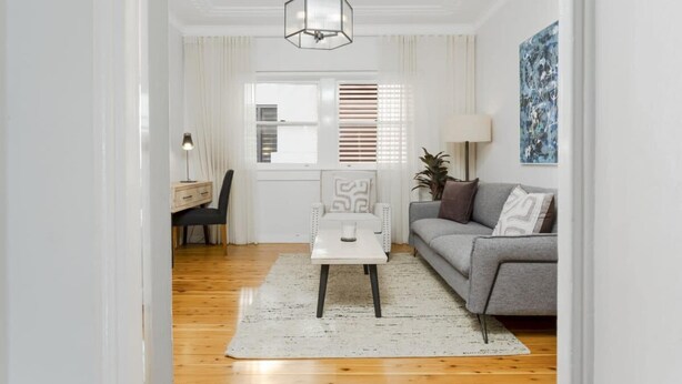 Una sala de estar moderna con un sofá gris, una mesa de centro blanca y piso de madera. Se ve un escritorio con una silla y una lámpara al fondo.