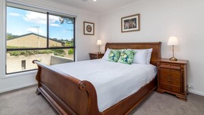 A bedroom with a wooden bed, two bedside tables with lamps, a ceiling fan, and a large window offering a view of a house and greenery. - Spacious and Restful Holiday Home with Backyard (Cook)