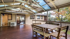 An open-plan living area with a wooden deck, a ceiling fan, and a view of a brick house through large windows. - Spacious and Restful Holiday Home with Backyard (Cook)