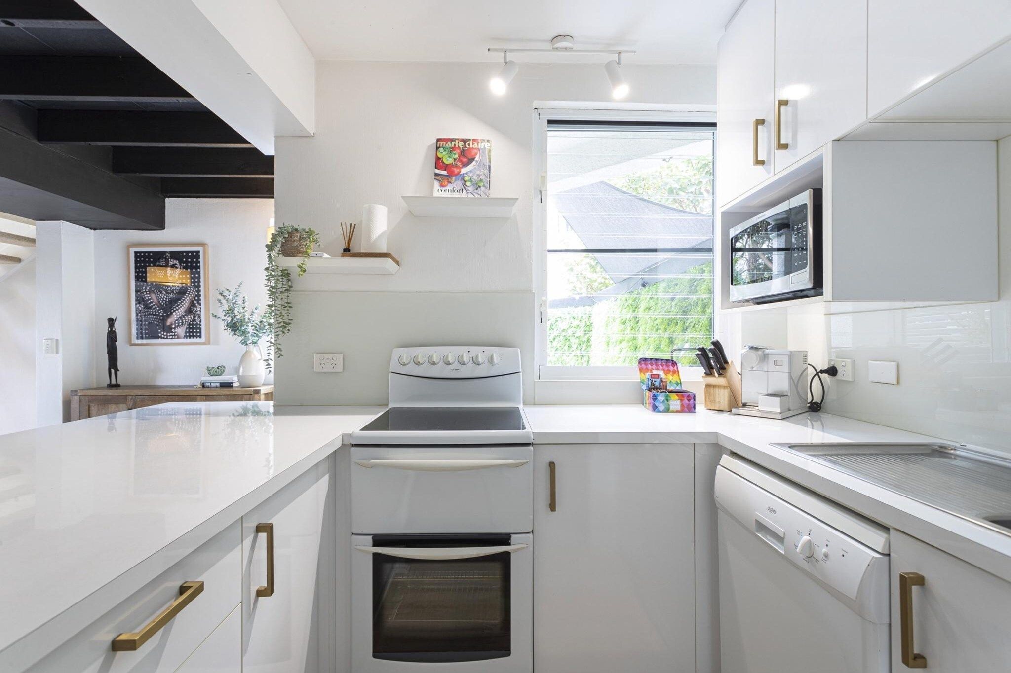 A modern kitchen with white cabinetry, a built-in oven, microwave, and dishwasher. There is a window providing natural light and a view of greenery outside.