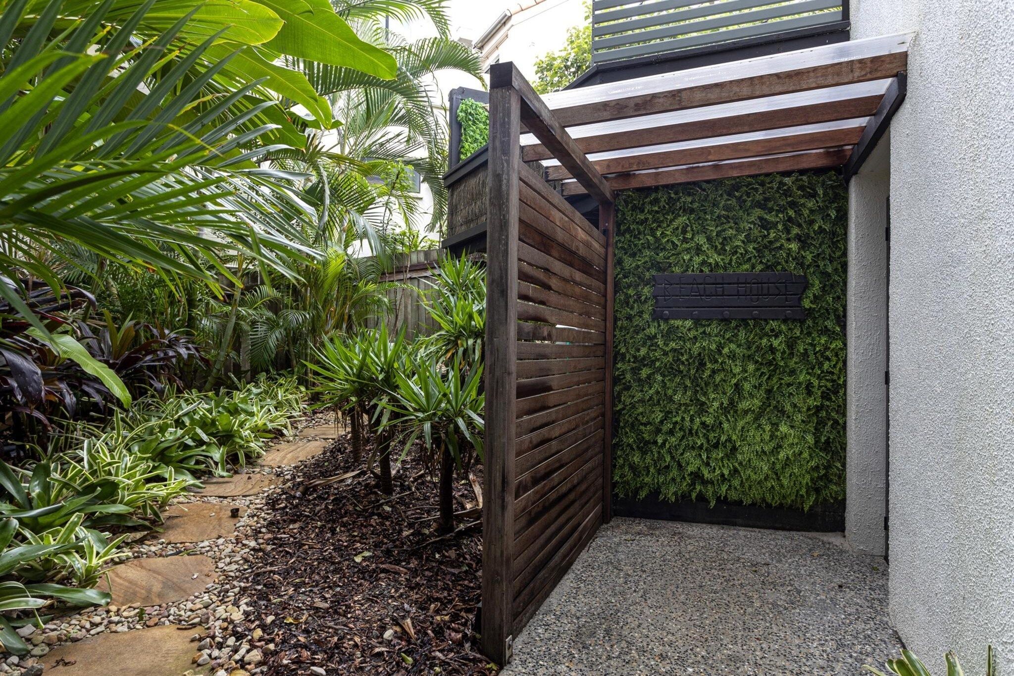 A gated entrance with a wooden gate, a paved pathway, and a variety of tropical plants.