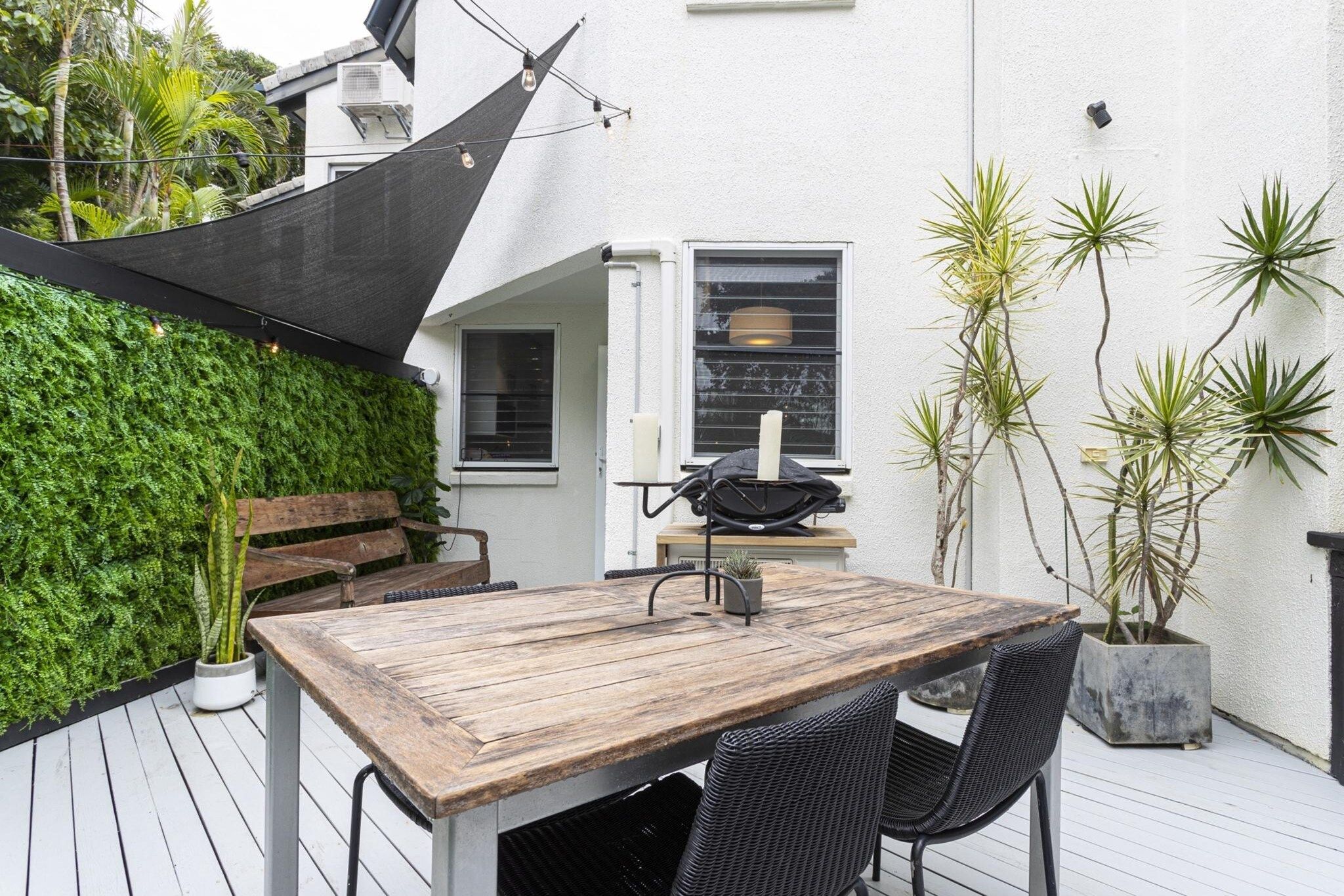 Une table en bois avec des chaises, un barbecue noir et un mur blanc agrémenté d’un jardin vertical vert.