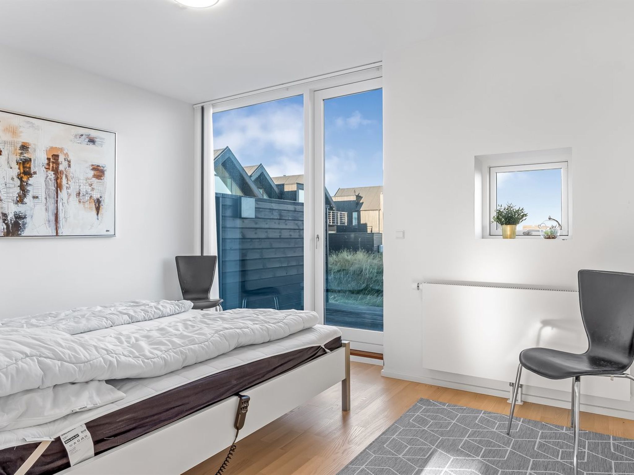 Ein modernes Schlafzimmer mit Bett, Sessel, Heizkörper und Fenster mit Blick auf Gebäude und Himmel.