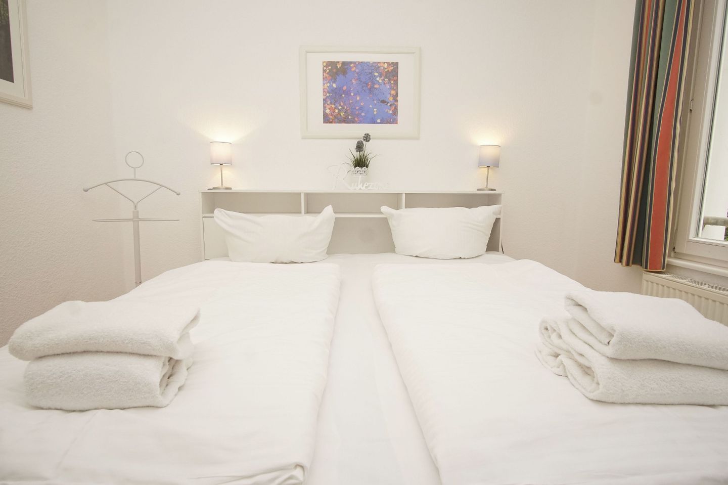 A hotel room with a double bed, two bedside tables, a wall-mounted artwork, and a window with curtains.