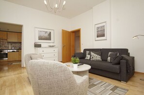 A living room with a grey sofa, a white coffee table, and a grey armchair. There is a kitchen in the background with wooden cabinets and a built-in oven. - Villa Odin - Beautiful 2-room apartment with a veranda - Villa Odin FeWo Ostseerauschen (Sellin)
