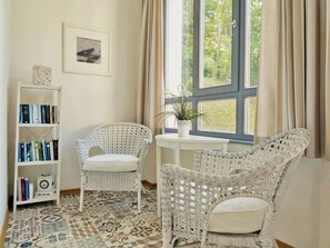 A room with a white wicker chair, a small white table with a plant, a bookshelf with books, and a patterned floor. - Villa Odin - beautiful 2-room apartment with veranda - Villa Odin FeWo Ostseerauschen (Sellin)