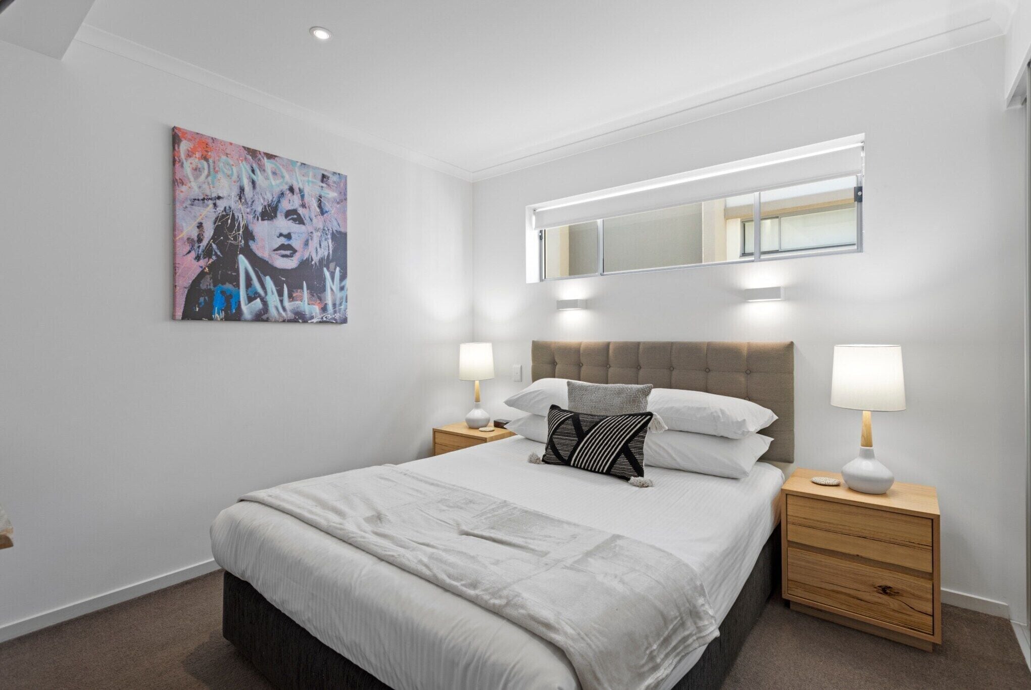 Un dormitorio moderno con una cama grande, dos mesitas de noche, un cabecero, una lámpara de techo, un espejo montado en la pared y una obra de arte.