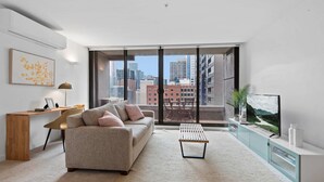 A modern living room with a sofa, coffee table, and TV, featuring large windows with a city view. - Modern 1-Bed City Apartment with Pool and Gym (Melbourne)