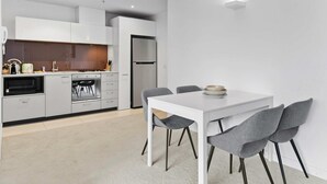 A modern kitchen with white cabinets, a built-in oven, and a refrigerator. There is a dining table with four grey chairs and a white tablecloth. - Modern 1-Bed City Apartment with Pool and Gym (Melbourne)