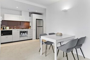 Eine moderne Küche mit weißen Schränken, einem eingebauten Backofen und einem Kühlschrank. Ein Esstisch mit vier grauen Stühlen und einer weißen Tischdecke ist ebenfalls vorhanden.