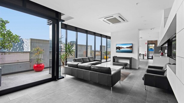 A modern living room with a grey sectional sofa, a red potted plant, and a large window offering a city view. - Modern 1-Bed City Apartment with Pool and Gym (Melbourne)