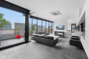 A modern living room with a grey sectional sofa, a red potted plant, and a large window offering a city view. - Modern 1-Bed City Apartment with Pool and Gym (Melbourne)