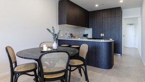 A modern kitchen with dark cabinetry, a central island, and a dining area with wicker chairs and a marble table. - Elegant 2-Bed Apartment with Incredible Bay Views (Batemans Bay)