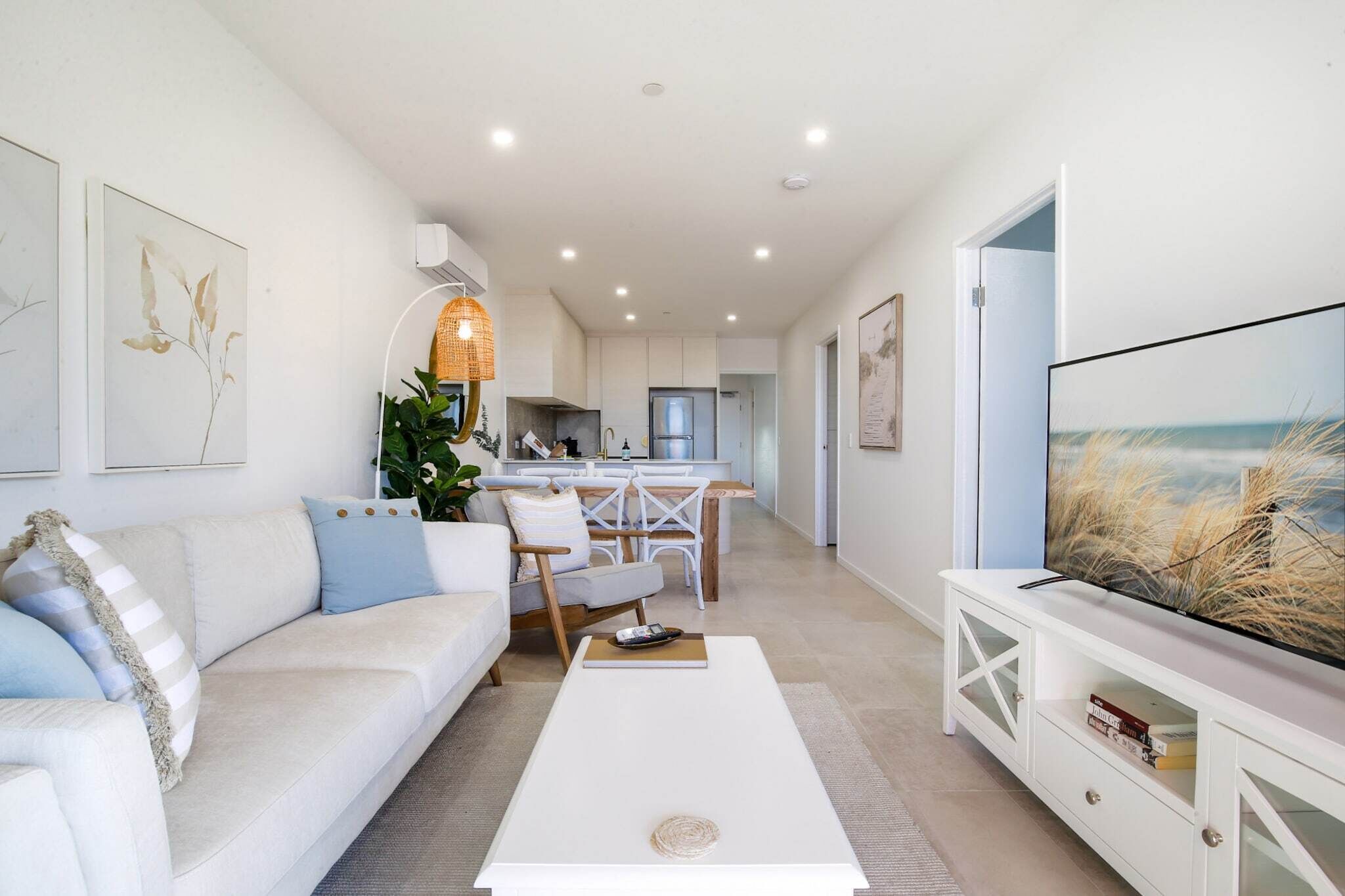 Un salon moderne avec un canapé blanc, une table basse et un téléviseur posé sur un meuble blanc. Au fond, on aperçoit une cuisine avec des meubles blancs et un coin repas comprenant une table et des chaises.