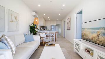 Un salon moderne avec un canapé blanc, une table basse et un téléviseur posé sur un meuble blanc. Au fond, on aperçoit une cuisine avec des meubles blancs et un coin repas comprenant une table et des chaises.