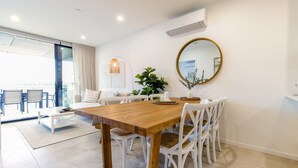 A modern dining area with a wooden table and white chairs, a round mirror, and a potted plant. - Bay View 2-Bedroom Apartment with BBQ (Batemans Bay)