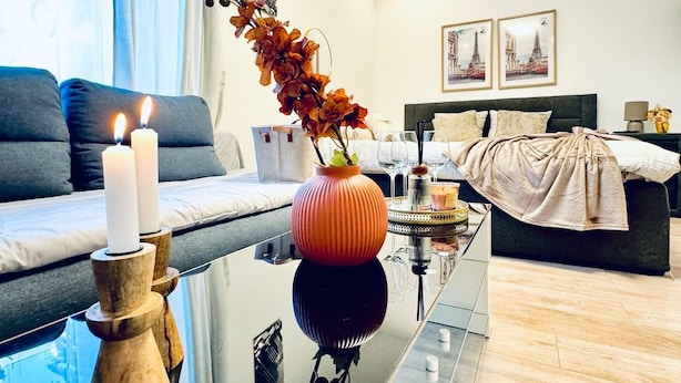 Een moderne woonkamer met een glazen salontafel, een bank, een bed en een vaas met bloemen.