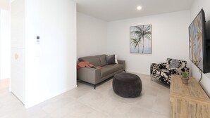 Uma sala de estar moderna com um sofá cinzento, um sofá estampado em preto e branco, um banquinho redondo e uma mesa de centro de madeira com uma planta.
