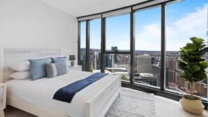 Ein modernes Schlafzimmer mit einem großen Bett, einem Nachttisch, einer Lampe und Blick auf eine Stadtlandschaft durch große Fenster.