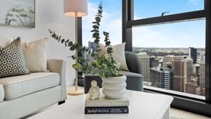 Ein modernes Wohnzimmer mit Blick auf eine Stadtlandschaft durch große Fenster, ein weißes Sofa, ein Couchtisch mit Büchern und eine Stehlampe.