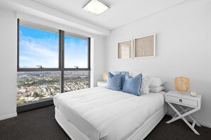 Ein modernes Hotelzimmer mit großem Fenster und Blick auf die Stadt, einem ordentlich bezogenen Bett, einem Nachttisch mit Lampe sowie zwei gerahmten Kunstwerken an der Wand.