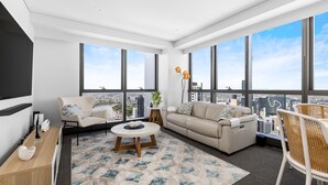 Ein modernes Wohnzimmer mit Sofa, Sessel und Couchtisch, großen Fenstern mit Blick auf die Stadt sowie einer Zierpflanze.